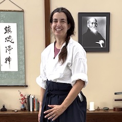 Adi Elkin, a woman with long dark hair in a braid, wearing a martial arts top and dark hakama, standing, smiling, in a room with a calligraphy scroll and a framed black-and-white portrait Ueshiba Morihei on the wall behind here.